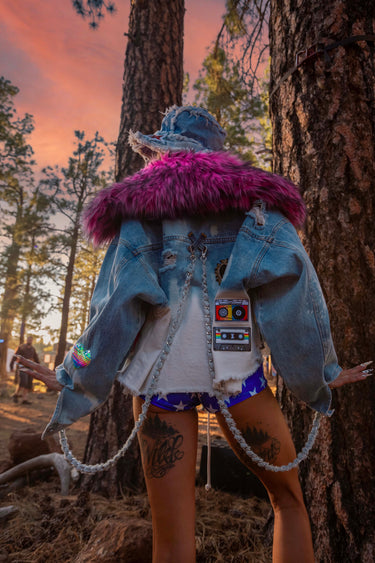 Back View of Model wearing Mixtape Oversized Denim Cropped Jacket with Love Sequin Cassettes and Heart Patches and Oversized 70's Style Pink Faux Fur Collar and Removable Hanging Denim Chain Straps | Festival Girly Rave Retro Style Fashion | Neon Nomade worn by Alyssa Risley in the forest at Desert Hearts